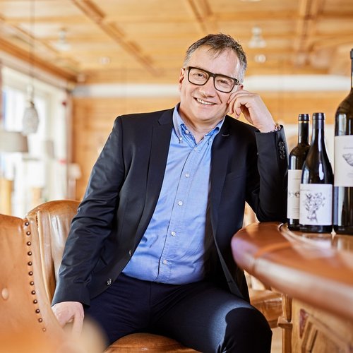 Hotel Sonnenhof Man with glasses in suit sitting at bar with wine bottles in bright room.