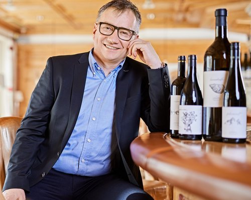Hotel Sonnenhof Mann mit Brille in Anzug sitzt an Bar mit Weinflaschen im hellen Raum.