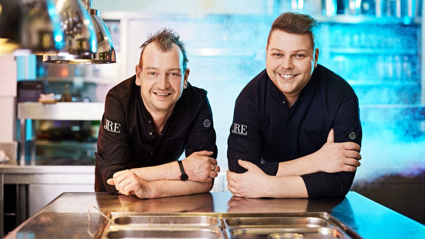 Hotel Sonnenhof Zwei lächelnde Köche in schwarzen Jacken lehnen an einem Küchentresen in einer Profiküche