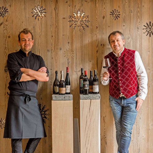 Hotel Sonnenhof Two men, one chef in black, one with wine glass, standing by wine bottles and wooden wall
