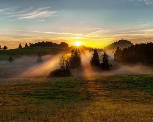 Hotel Sonnenhof Sonnenaufgang über nebligen Hügeln mit Bäumen und klar blauem Himmel.