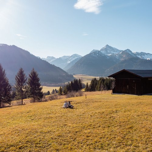 Hotel Sonnenhof Zwei Personen sitzen auf einer Wiese neben einer Hütte mit Blick auf schneebedeckte Berge