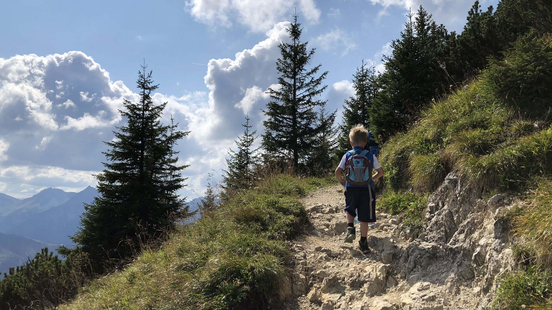 Hotel Sonnenhof Kind wandert auf felsigem Bergweg mit Kiefern und bewölktem Himmel