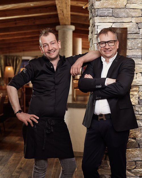 Hotel Sonnenhof Two smiling men, one in chef attire, the other in a suit, leaning against a stone wall