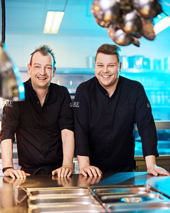 Patrick_Mueller_Fotocredit_Guenter_Standl_web_sonnenhof_0225-0399.jpg Two smiling chefs in black jackets in a modern kitchen setting