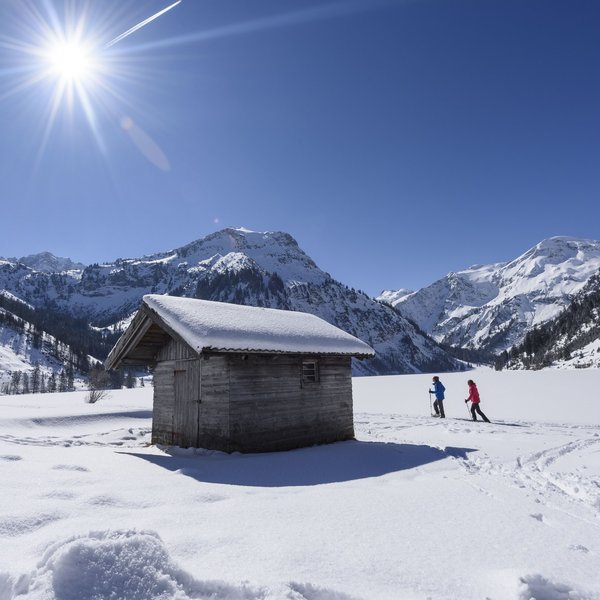 Hotel Sonnenhof Schneebedeckte Hütte mit zwei Skifahrern und Bergen unter strahlend blauem Himmel