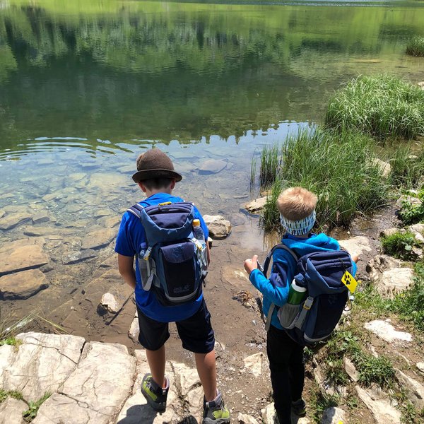 Hotel Sonnenhof Zwei Kinder mit Rucksäcken am Ufer eines klaren Bergsees