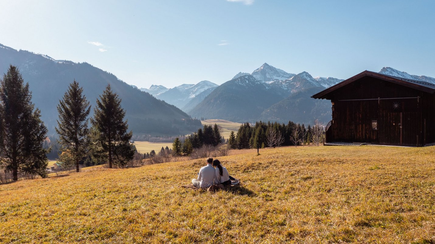 Hotel Sonnenhof Paar sitzt auf einer Wiese vor einer Berghütte mit Alpen im Hintergrund