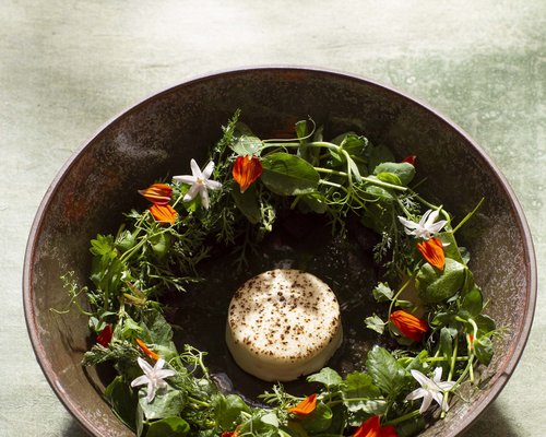Hotel Sonnenhof Käse in der Mitte, umgeben von grünen Kräutern und essbaren Blüten in einer braunen Schüssel