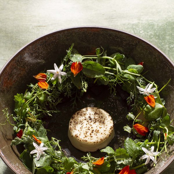 Hotel Sonnenhof Käse in der Mitte, umgeben von grünen Kräutern und essbaren Blüten in einer braunen Schüssel