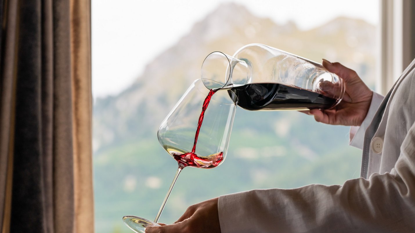 Hotel Sonnenhof Person gießt Rotwein aus Karaffe in Weinglas vor Bergkulisse